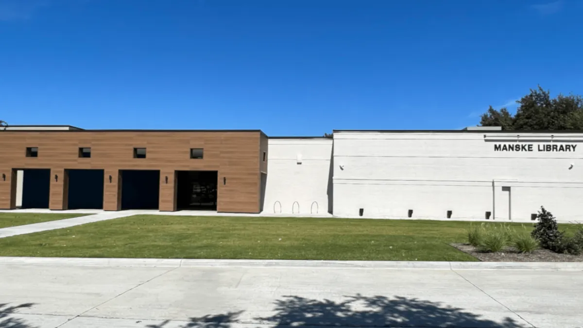 A photograph of the library from the street: a timber-clad entrance block on the left, a white masonry wall carrying the words MANSKE LIBRARY on the right, and an empty lawn in front.